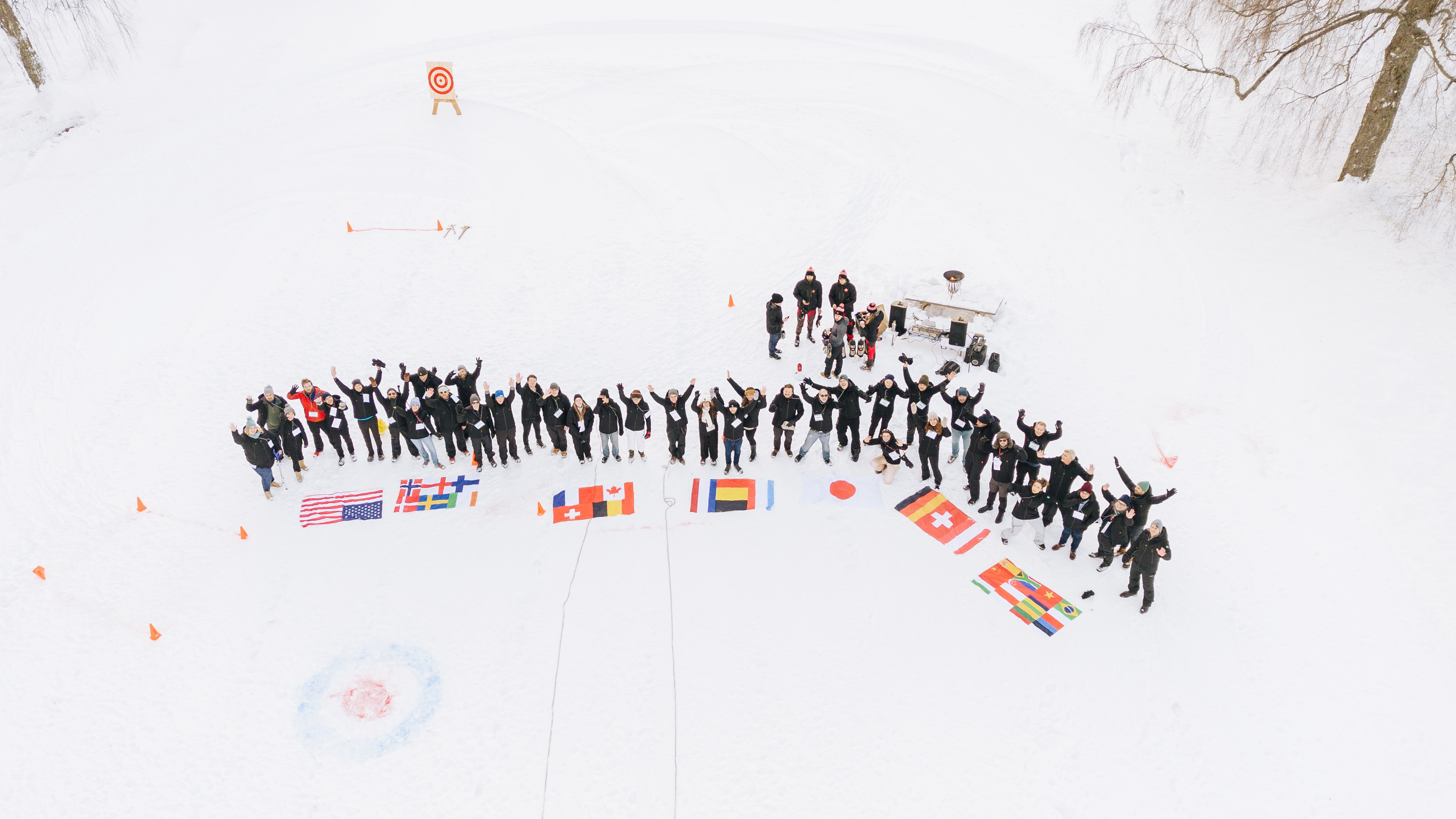 Aerial drone view of the Catenda team forming an arrow shape on snow, arms raised, with colourful international flags laid out in front of them