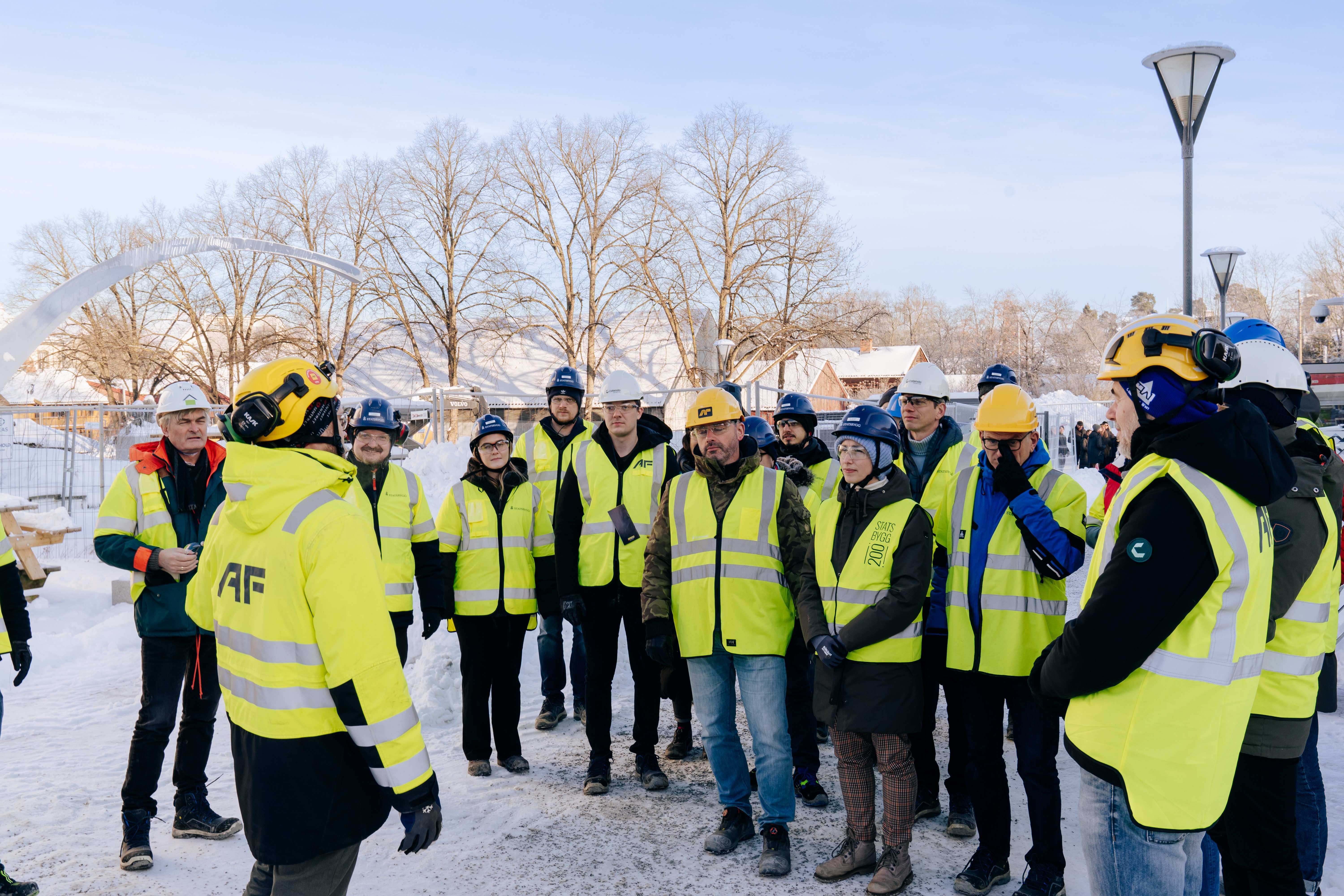 Large group of Catenda team members in yellow hi-vis vests listening to a briefing at a snowy construction site under blue sky