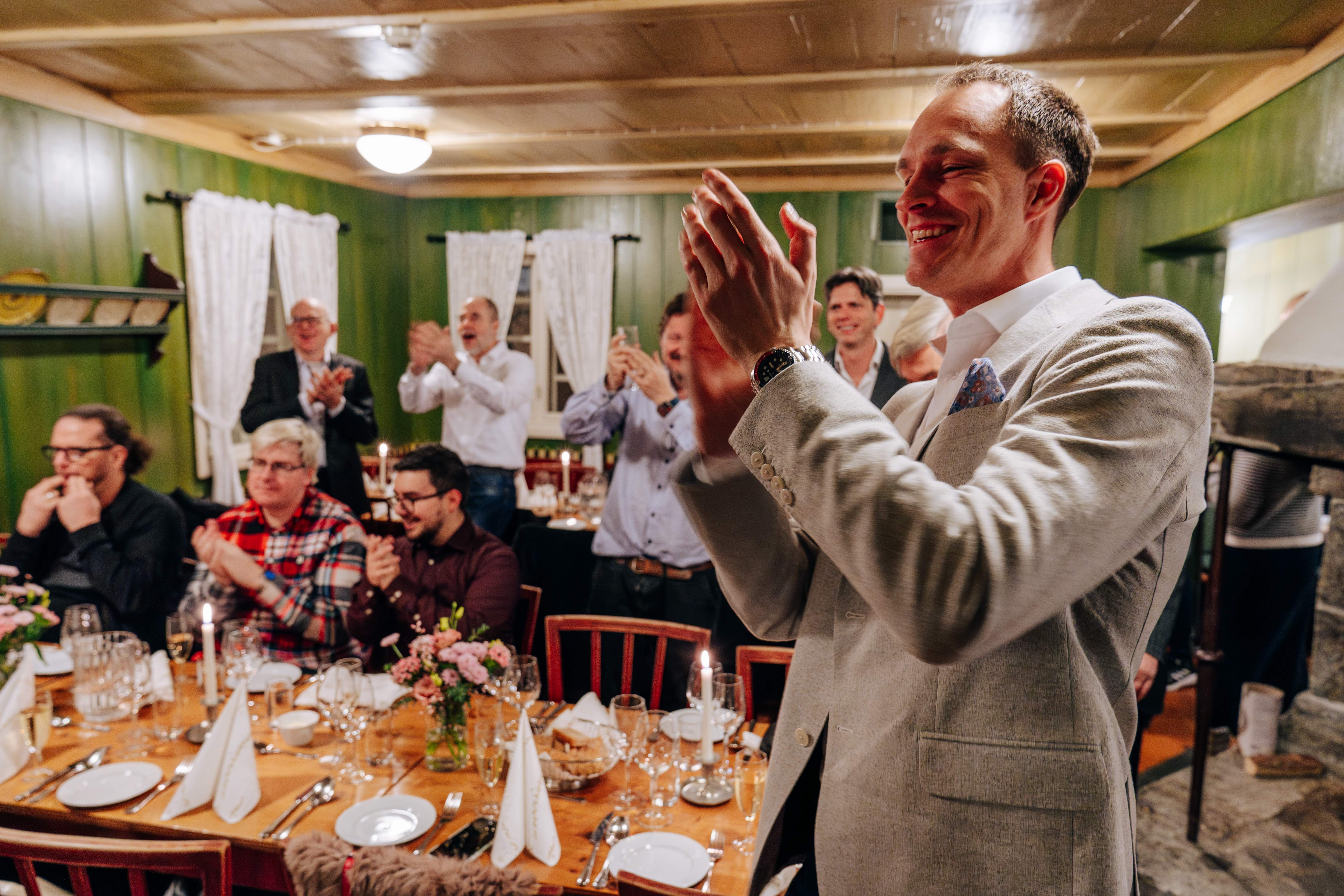 Catenda team celebrating at a candlelit dinner inside a cosy cabin with green wooden walls, a man in a grey suit applauding