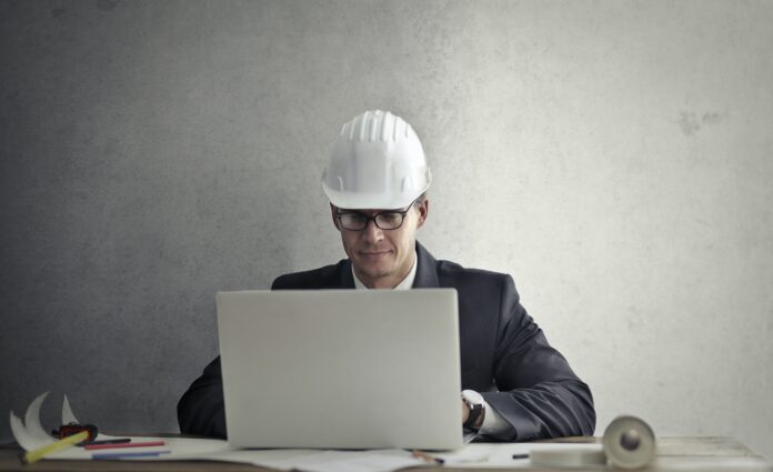 construction worker with white helmet sitting on table with a laptop and some architectural plans
