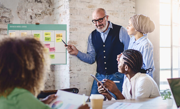 A senior man with a white beard and glasses points at a business model canvas filled with sticky notes. Beside him, a Middle Eastern woman with a turban, and colleagues taking notes.