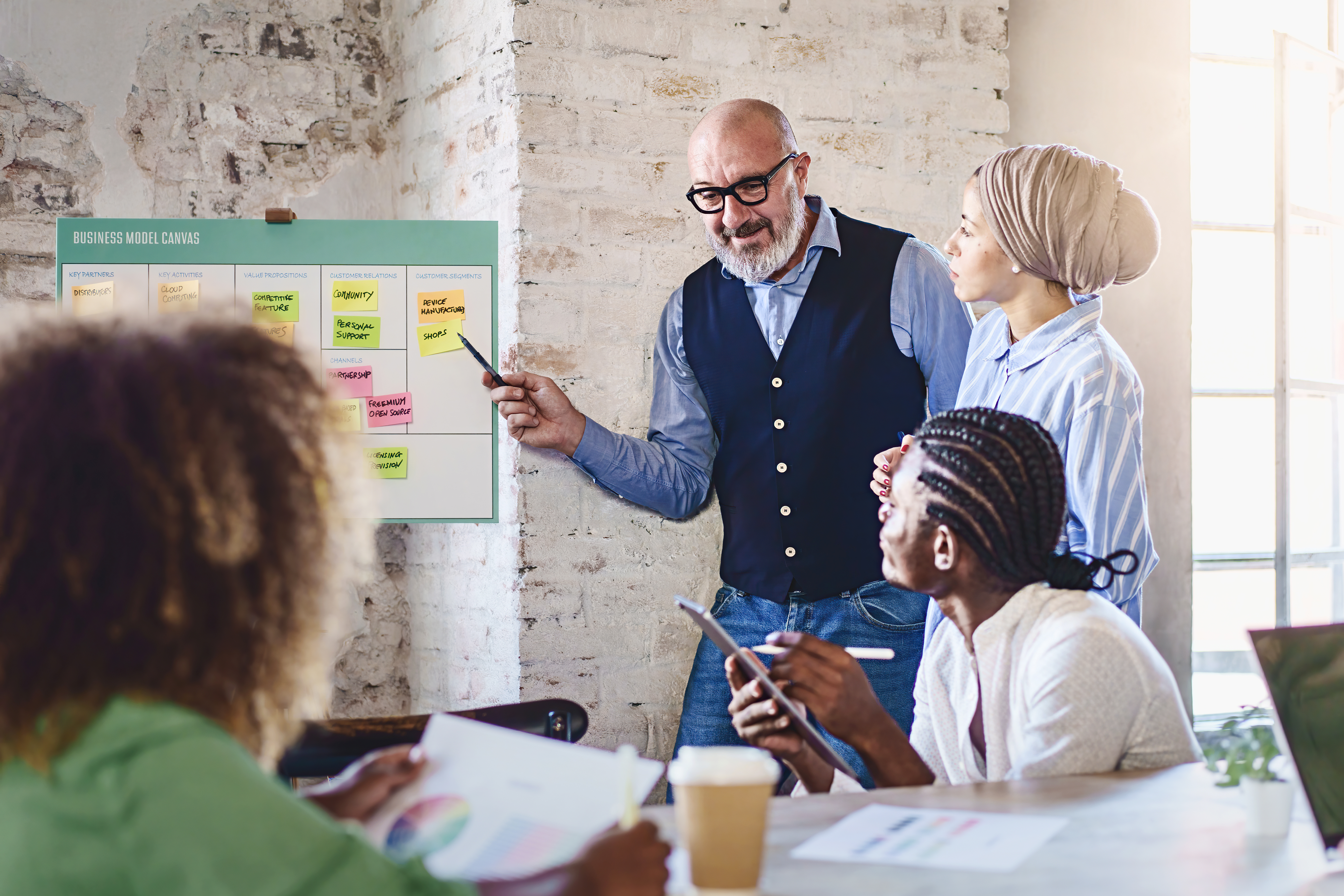 A senior man with a white beard and glasses points at a business model canvas filled with sticky notes. Beside him, a Middle Eastern woman with a turban, and colleagues taking notes.