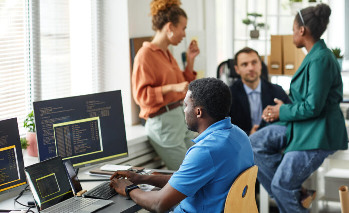 African IT specialist working with software typing on keyboard sitting at table with computers while his colleagues working in team in background