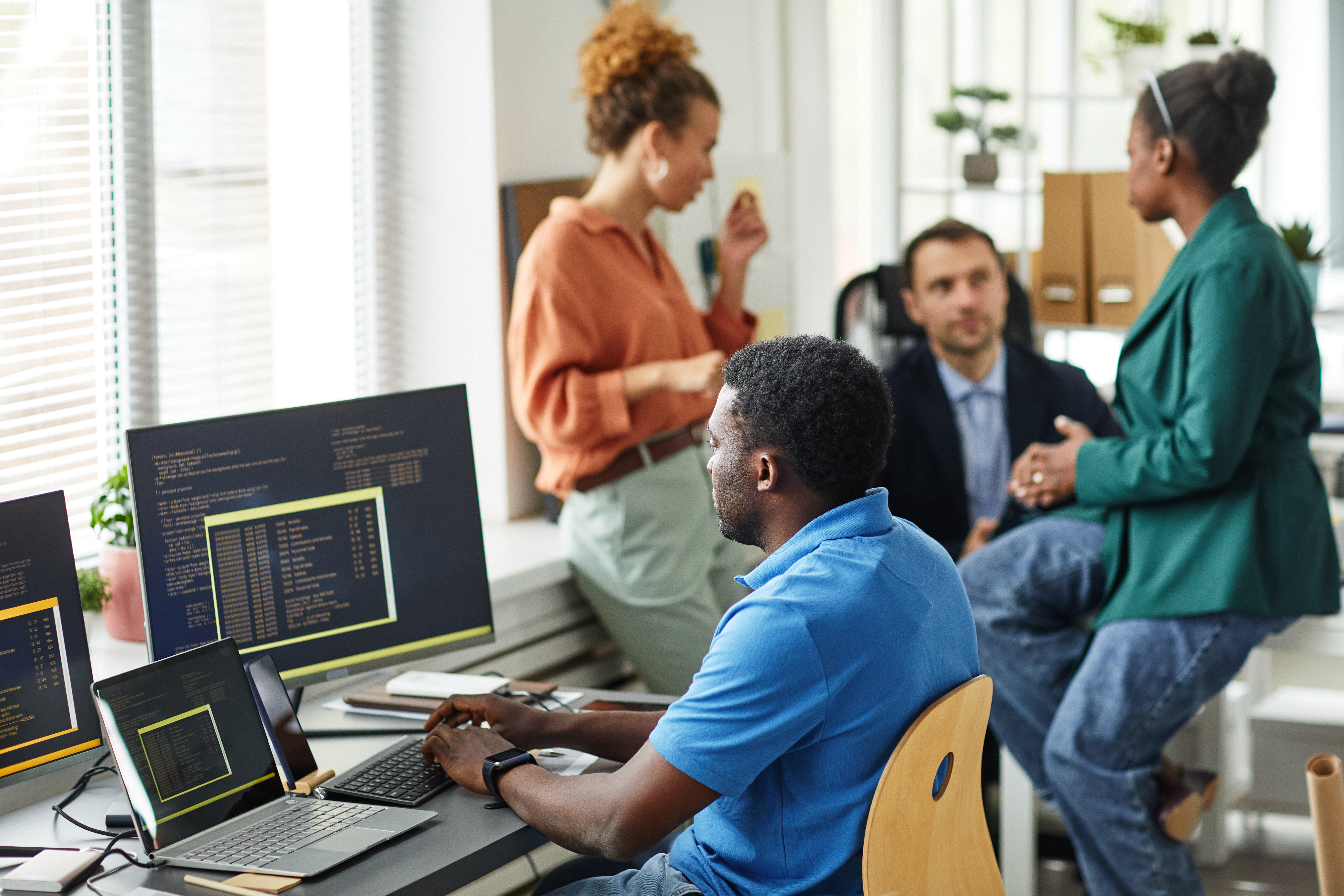 African IT specialist working with software typing on keyboard sitting at table with computers while his colleagues working in team in background