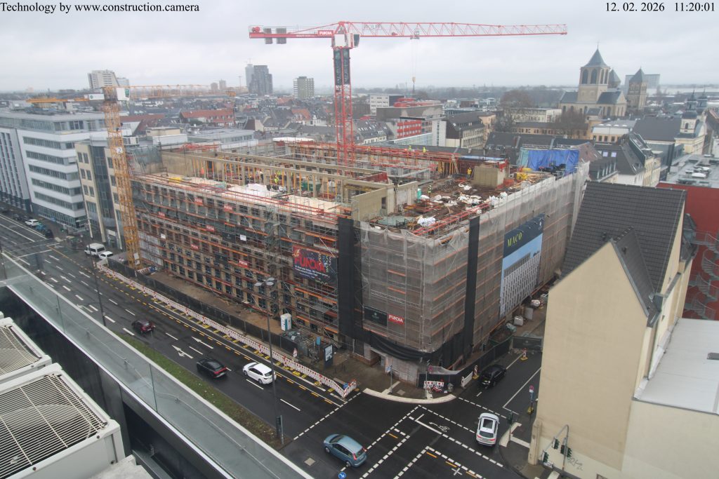 Aerial construction site view of the MACO building in February 2026, showing scaffolding, a red tower crane, and the building's structural frame.