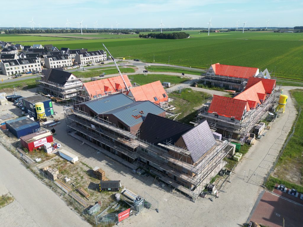 Aerial drone photo of a residential construction site in Zeewolde with multiple building blocks under construction, scaffolding, and wind turbines in the background
